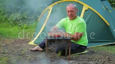 一个穿着绿色T恤的男人坐在森林里，靠着绿色的帐篷，把<strong>烧</strong>烤放在<strong>烧烤架</strong>上。 总体计划